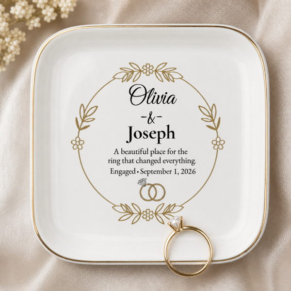 A square white ceramic ring holder with rounded corners, featuring a gold border and personalized printed text and floral wreath design. A gold ring rests in one of the holder's corners on a beige satin fabric background with small pearl flowers in the corner.