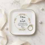 A square white ceramic ring box with rounded corners and a gold rim, printed with a floral wreath and personalized names 'Olivia & Joseph' with wedding text. A gold ring with a clear gemstone rests inside the box.