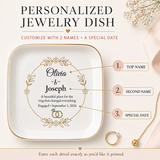 A square white personalized jewelry dish with gold trim and printed custom text and ring illustration, shown on a soft beige background with decorative flowers, a gold ring, and callout labels pointing to top name, second name, and special date.