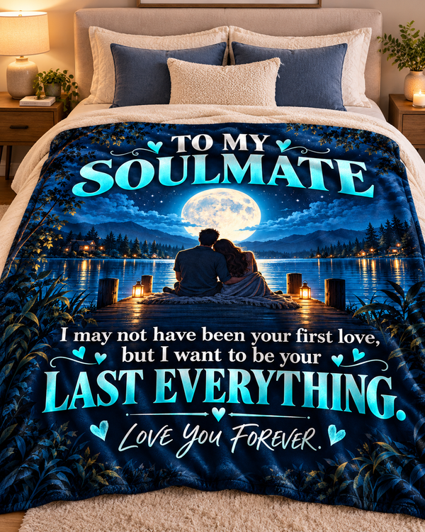 A bed with a printed comforter or blanket featuring a romantic night scene: two people sitting on a dock by a lake under a large full moon, with the text 'TO MY SOULMATE' and a sentimental message below in bright teal and white lettering.