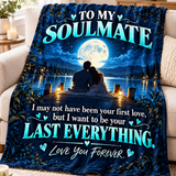 A printed blanket draped over a beige sofa in a living room. The blanket features a dark blue night-sky scene with a couple sitting by a lake at a dock, a large full moon, and large turquoise text that reads, 'TO MY SOULMATE' and 'I may not have been your first love, but I want to be your LAST EVERYTHING.