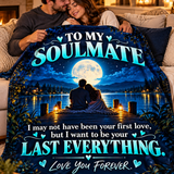A couple huddled under a cozy blanket on a couch in a warmly lit living room. The blanket features a printed night scene with a couple sitting by a lake under a full moon, with the text 'TO MY SOULMATE I may not have been your first love, but I want to be your LAST EVERYTHING.