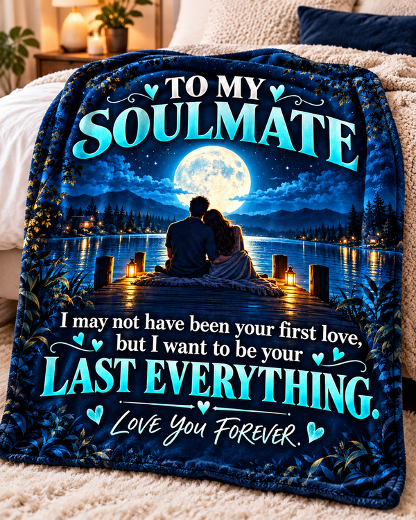 A dark blue fleece blanket draped over a bed, featuring a printed graphic of a couple sitting by a lake at night under a full moon, with the text 'TO MY SOULMATE I may not have been your first love, but I want to be your LAST EVERYTHING. Love You Forever.