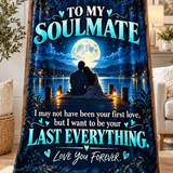 A printed blanket or throw draped over a wooden stand in a living room setting. The design shows a couple sitting on a dock at night by a lake under a full moon, with large teal text reading 'TO MY SOULMATE' and 'I may not have been your first love, but I want to be your LAST EVERYTHING.