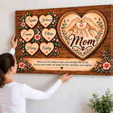 A woman holds a large personalized wooden canvas print against a white wall. The artwork features a heart-shaped design with hands forming a heart, the word 'Mom' in script, six individual heart names with small red hearts, floral details, and a sentimental message for Mom at the bottom.