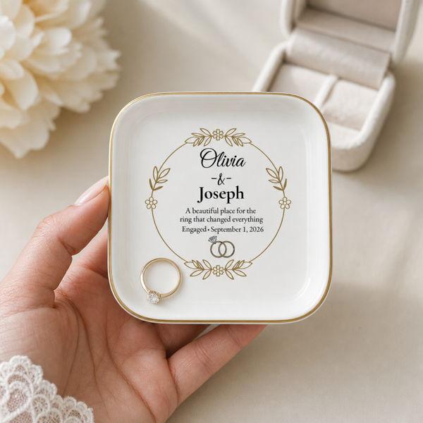 A hand holds a small square white ring box with rounded corners and gold trim. The top is printed with a personalized floral wreath design and the names Olivia & Joseph with a small engagement date.